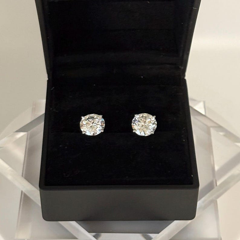 May include: A pair of sparkling diamond stud earrings displayed in a black jewelry box. The earrings are round-cut and set against a black velvet interior, creating a striking contrast. The box is open, revealing the brilliance of the gemstones.