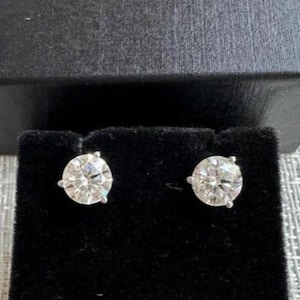 May include: A pair of diamond stud earrings. Each earring features a round, brilliant-cut diamond set in a silver or white gold prong setting. The earrings are displayed on a black velvet surface, with a dark gray box in the background.