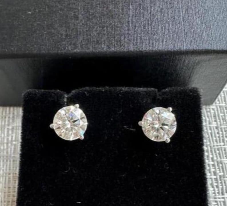 May include: A pair of diamond stud earrings. Each earring features a round, brilliant-cut diamond set in a silver or white gold prong setting. The earrings are displayed on a black velvet surface, with a dark gray box in the background.