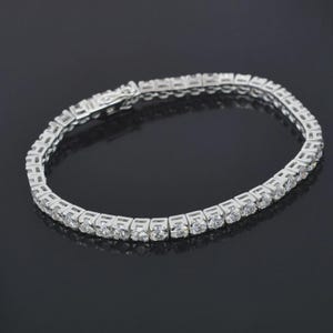 May include: A silver tennis bracelet with multiple round, clear gemstones set in a row. The bracelet is displayed on a dark surface, reflecting the light from the gemstones.