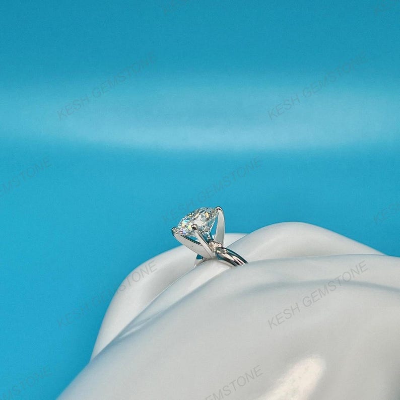 Op de afbeelding: Een zilveren verlovingsring met een grote, heldere, rond geslepen edelsteen. De ring ligt op een wit, gebogen oppervlak tegen een helderblauwe achtergrond. Het ontwerp van de ring is klassiek en elegant.
