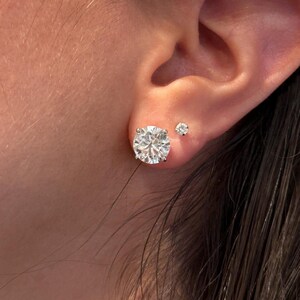 May include: Close-up of a person's ear with two diamond stud earrings. The larger earring is a round-cut diamond, and the smaller earring is a smaller round-cut diamond. The earrings are set in a silver or white gold setting.