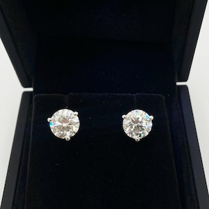 May include: A pair of round-cut diamond stud earrings displayed in an open black jewelry box. The diamonds are set in a silver or white gold prong setting. The earrings are sparkling and reflect light.