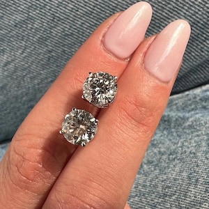 May include: Two round-cut diamond stud earrings. The diamonds are set in a silver metal. The earrings are held up by a person's fingers with pink nail polish, against a denim background.