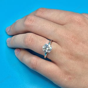 Op de afbeelding: Een zilveren ring met een grote, ronde, heldere edelsteen. De ring heeft een eenvoudige band. De ring wordt getoond op een hand tegen een blauwe achtergrond.