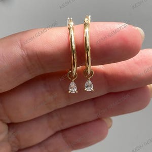 May include: A pair of gold hoop earrings with a teardrop-shaped, clear gemstone pendant. The earrings are displayed against a neutral background. The earrings are a classic jewelry item.