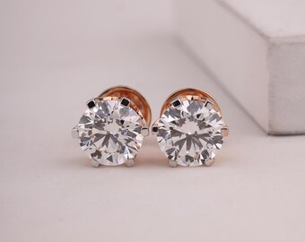 May include: A pair of diamond stud earrings. Each earring features a large, round, brilliant-cut diamond set in a rose gold and silver setting. The diamonds sparkle with numerous facets, reflecting light. The earrings are displayed on a white surface.