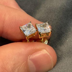 May include: A pair of square-cut, clear gemstone stud earrings with gold-colored settings. The earrings are held between fingers, showcasing their sparkling facets. The gold setting adds a touch of elegance to the jewelry.