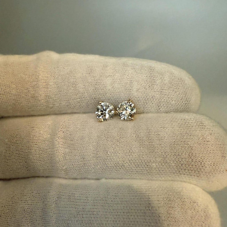 May include: A pair of diamond stud earrings. Each earring features a round, brilliant-cut diamond set in a gold prong setting. The diamonds sparkle against the neutral background, showcasing their clarity and brilliance. A classic jewelry item.