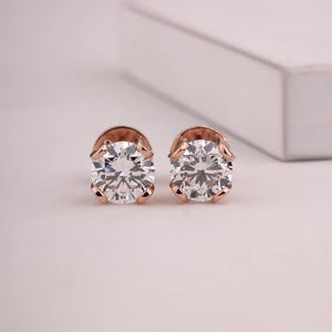 Könnte beinhalten: Ein Paar roségoldene Ohrstecker mit großen, klaren, rund geschliffenen Edelsteinen. Die Ohrringe sind in einer Vier-Zacken-Fassung gefasst, die die Brillanz der Steine hervorhebt. Der Hintergrund ist eine weiche, neutrale Farbe.