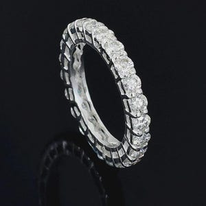May include: A silver eternity ring with a continuous row of round-cut diamonds. The ring's band has a detailed, textured design. The diamonds sparkle against the dark background, creating a luxurious and elegant look.