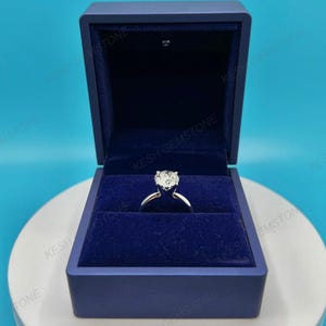 Op de afbeelding: Een diamanten verlovingsring met een grote, heldere edelsteen wordt getoond in een donkerblauwe, met fluweel gevoerde doos. De ring heeft een zilveren band. De doos is open en toont de ring tegen een donkerblauwe achtergrond.