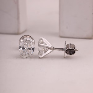 May include: Two diamond stud earrings. One earring features an oval-cut diamond, while the other showcases a round-cut diamond. Both are set in a silver metal. The earrings are displayed on a white surface.
