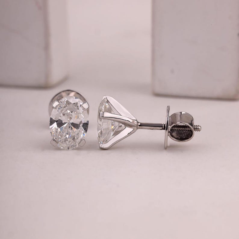 May include: Two diamond stud earrings. One earring features an oval-cut diamond, while the other showcases a round-cut diamond. Both are set in a silver metal. The earrings are displayed on a white surface.