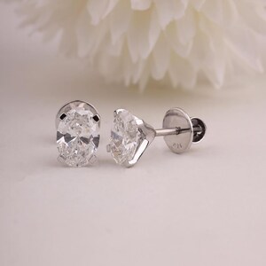 May include: A pair of diamond stud earrings. Each earring has a clear, oval-cut diamond set in a silver metal setting. The earrings are displayed on a white surface, with a blurred white flower in the background.