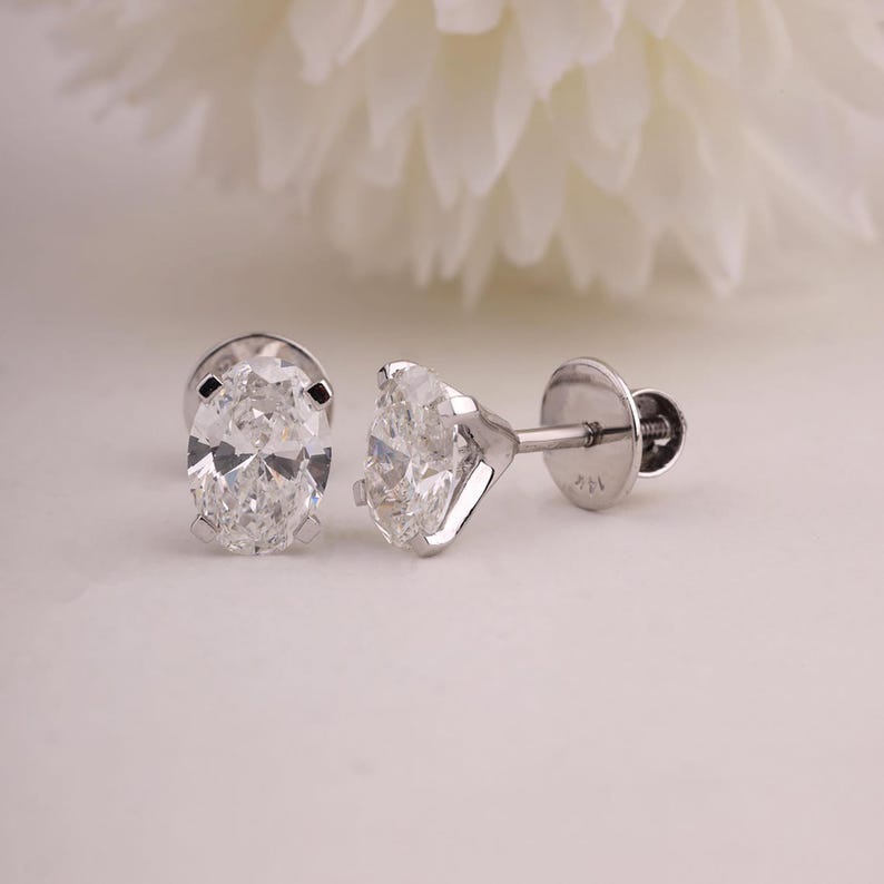 May include: A pair of diamond stud earrings. Each earring has a clear, oval-cut diamond set in a silver metal setting. The earrings are displayed on a white surface, with a blurred white flower in the background.