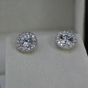 May include: A pair of diamond stud earrings. Each earring features a large, round, clear diamond surrounded by a halo of smaller diamonds, set in a silver or white gold setting. The earrings are displayed in a white jewelry box.