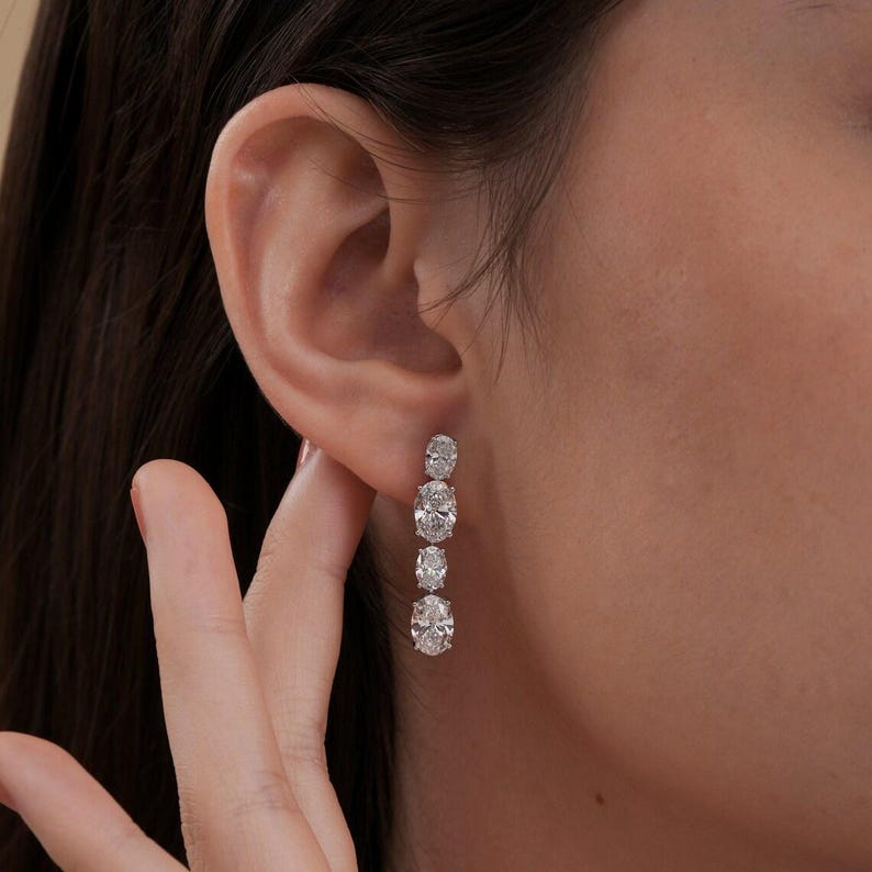 May include: Elegant drop earrings featuring four oval-cut diamonds. The diamonds are set in a linear arrangement, creating a sparkling and sophisticated look. The earrings are shown on a person's ear, highlighting their design and brilliance.