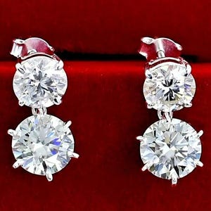 May include: A pair of silver drop earrings with two large, round, clear gemstones. The earrings are set against a deep red background, highlighting the sparkle of the stones. The earrings have a classic, elegant design.