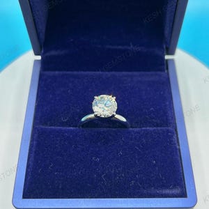 Op de afbeelding: Een zilveren verlovingsring met een grote, ronde, heldere edelsteen gezet in een vier-klauwzetting. De ring wordt gepresenteerd in een donkerblauwe fluwelen ringdoos. De doos is open en onthult de ring.