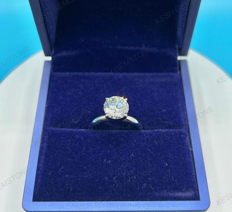 Op de afbeelding: Een zilveren verlovingsring met een grote, ronde, heldere edelsteen gezet in een vier-klauwzetting. De ring wordt gepresenteerd in een donkerblauwe fluwelen ringdoos. De doos is open en onthult de ring.