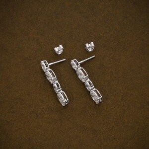 May include: A pair of silver drop earrings with multiple clear, rectangular-cut stones. The earrings are displayed with their matching earring backs on a brown surface. The stones are set in a linear design.