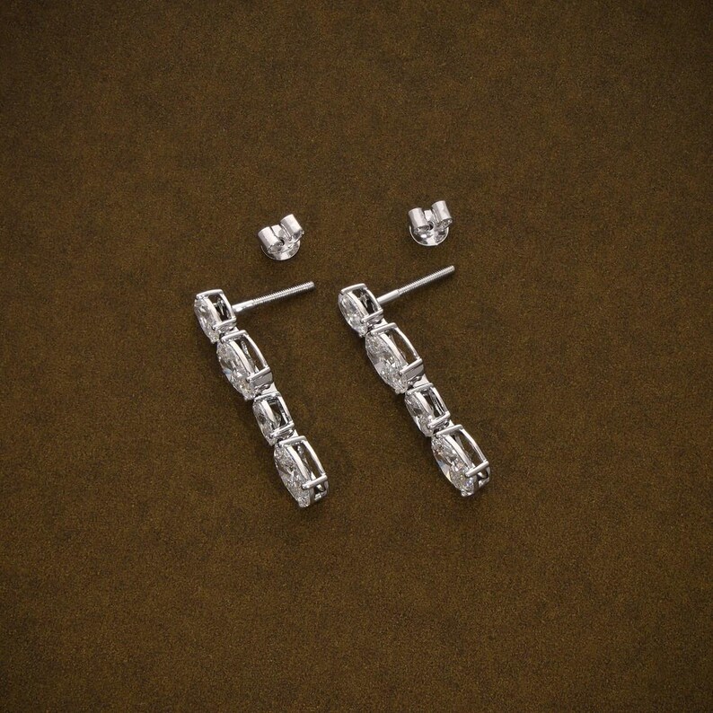May include: A pair of silver drop earrings with multiple clear, rectangular-cut stones. The earrings are displayed with their matching earring backs on a brown surface. The stones are set in a linear design.
