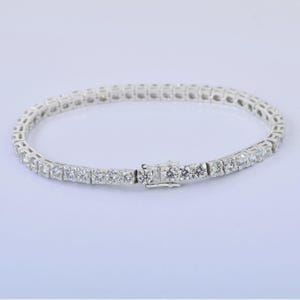 May include: A silver tennis bracelet with a row of round-cut, clear gemstones. The bracelet is clasped and lies on a white surface. The gemstones are evenly spaced and reflect light.