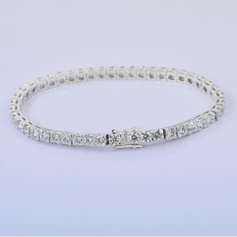 May include: A silver tennis bracelet with a row of round-cut, clear gemstones. The bracelet is clasped and lies on a white surface. The gemstones are evenly spaced and reflect light.