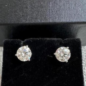 Puede incluir: Un par de pendientes de diamantes redondos y transparentes, colocados sobre una pantalla de terciopelo negro. Los pendientes están engastados en un engaste de plata u oro blanco, con tres garras que sujetan cada piedra.