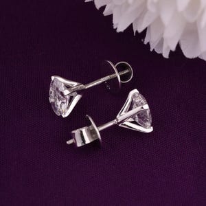 May include: A pair of diamond stud earrings. Each earring has a clear, round-cut diamond set in a silver metal prong setting. The earrings are displayed on a dark purple surface, with a white flower in the background.
