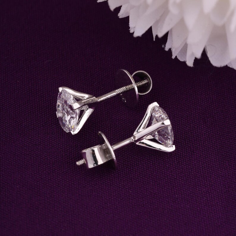 May include: A pair of diamond stud earrings. Each earring has a clear, round-cut diamond set in a silver metal prong setting. The earrings are displayed on a dark purple surface, with a white flower in the background.