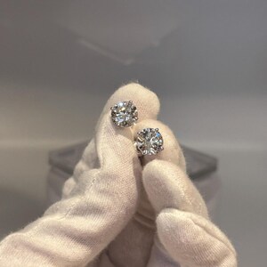 May include: A pair of sparkling diamond stud earrings. The round-cut diamonds are held in place by a silver setting. The earrings are held by a gloved hand against a soft, neutral background. The diamonds reflect light, creating a brilliant sparkle.