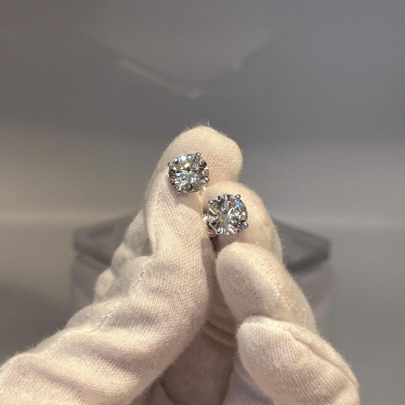 May include: A pair of sparkling diamond stud earrings. The round-cut diamonds are held in place by a silver setting. The earrings are held by a gloved hand against a soft, neutral background. The diamonds reflect light, creating a brilliant sparkle.