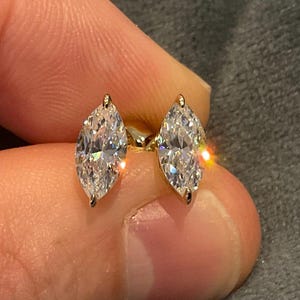 May include: A pair of marquise-cut diamond stud earrings. The diamonds are set in gold prongs and are held between fingers. The earrings sparkle with light, showcasing their facets. The background is a blurred gray.