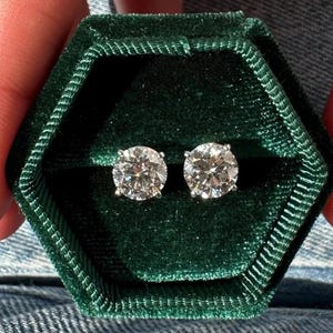 May include: A pair of diamond stud earrings displayed in a dark green velvet jewelry box. The earrings are round-cut, with a brilliant sparkle. The box is hexagonal, with a soft, plush texture, and is held by a person.