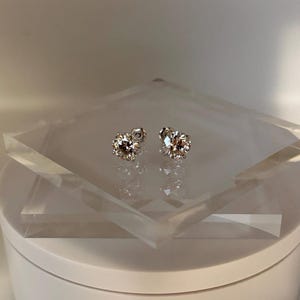 May include: A pair of sparkling stud earrings with a round, faceted design. The earrings are set on a clear, geometric display, showcasing their brilliance. The studs are a warm, champagne color, reflecting light.