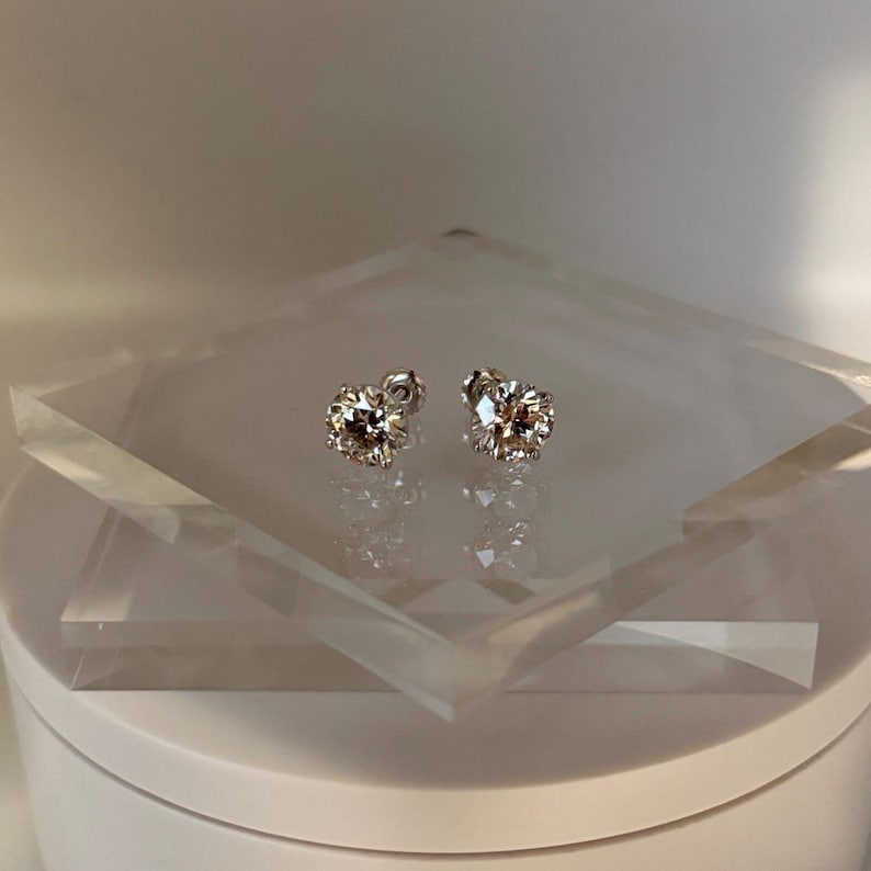 May include: A pair of sparkling stud earrings with a round, faceted design. The earrings are set on a clear, geometric display, showcasing their brilliance. The studs are a warm, champagne color, reflecting light.