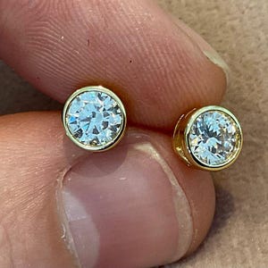 May include: A pair of gold bezel-set stud earrings. Each earring features a round, clear gemstone. The gold settings have a polished finish, and the gemstones sparkle. The earrings are shown close up.