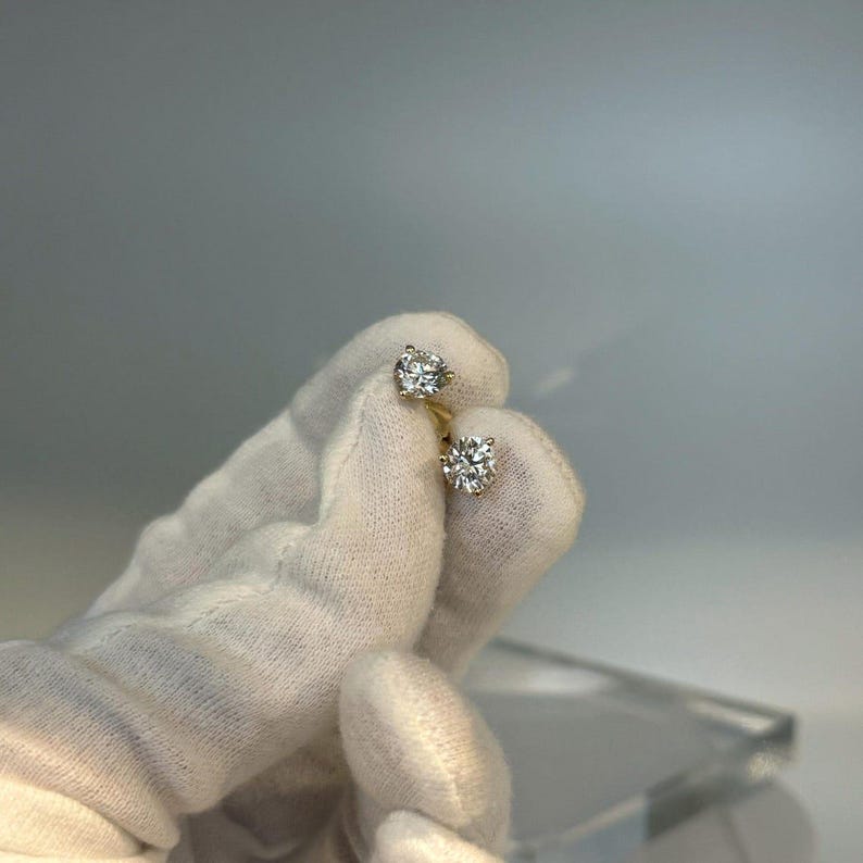 May include: A pair of diamond stud earrings held by a gloved hand. The earrings feature round-cut diamonds set in gold. The diamonds sparkle against the neutral background, showcasing their brilliance and clarity.