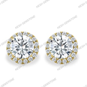May include: A pair of round, gold-colored stud earrings. Each earring features a large, clear, round-cut gemstone surrounded by a halo of smaller, clear gemstones. The earrings are set against a white background.