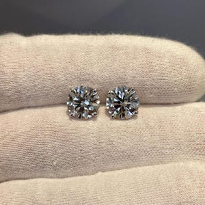 May include: A pair of round-cut diamond stud earrings. The earrings are set in a silver metal and are displayed on a white surface. The diamonds sparkle, reflecting light. The earrings are a classic jewelry item.
