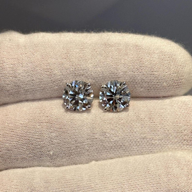 May include: A pair of round-cut diamond stud earrings. The earrings are set in a silver metal and are displayed on a white surface. The diamonds sparkle, reflecting light. The earrings are a classic jewelry item.