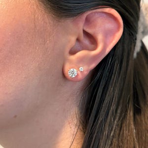 May include: A close-up of an ear with two diamond stud earrings. The larger earring is a round, brilliant-cut diamond, and the smaller earring is a smaller round diamond. The earrings are set in a silver or white gold setting.