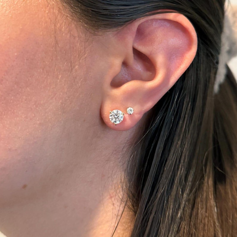 May include: A close-up of an ear with two diamond stud earrings. The larger earring is a round, brilliant-cut diamond, and the smaller earring is a smaller round diamond. The earrings are set in a silver or white gold setting.