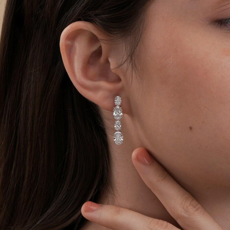 May include: A close-up of a dangling earring with four oval-shaped, sparkling gemstones. The earring is silver-toned and hangs vertically from the ear. The gemstones are clear and reflect light, creating a brilliant display. The earring is worn on a person's ear.