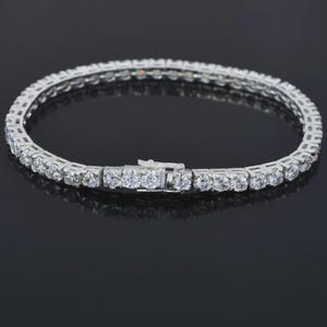 May include: A silver-toned tennis bracelet with a row of round, clear gemstones. The bracelet has a secure clasp and is displayed on a reflective black surface. The gemstones are evenly spaced and catch the light, creating a sparkling effect.