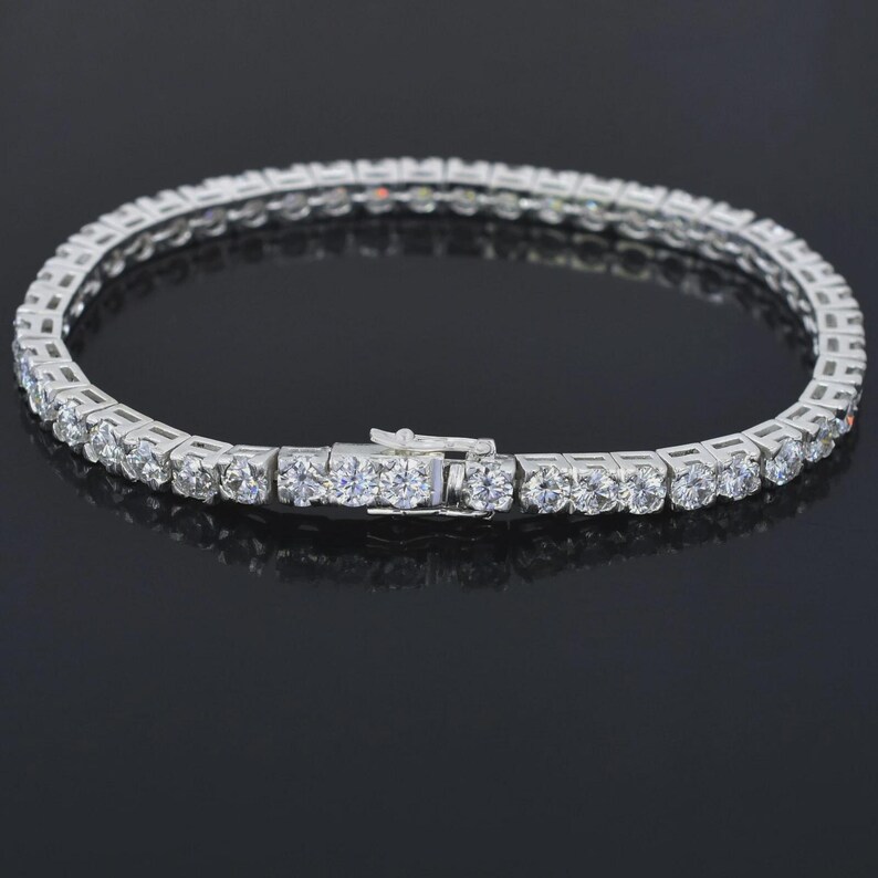 May include: A silver-toned tennis bracelet with a row of round, clear gemstones. The bracelet has a secure clasp and is displayed on a reflective black surface. The gemstones are evenly spaced and catch the light, creating a sparkling effect.