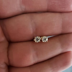 Pode incluir: Um par de brincos pequenos, redondos, com diamantes, em ouro. Os diamantes são claros e brilhantes, e os brincos são exibidos em um fundo neutro. Estes brincos são um item de joalheria clássico.