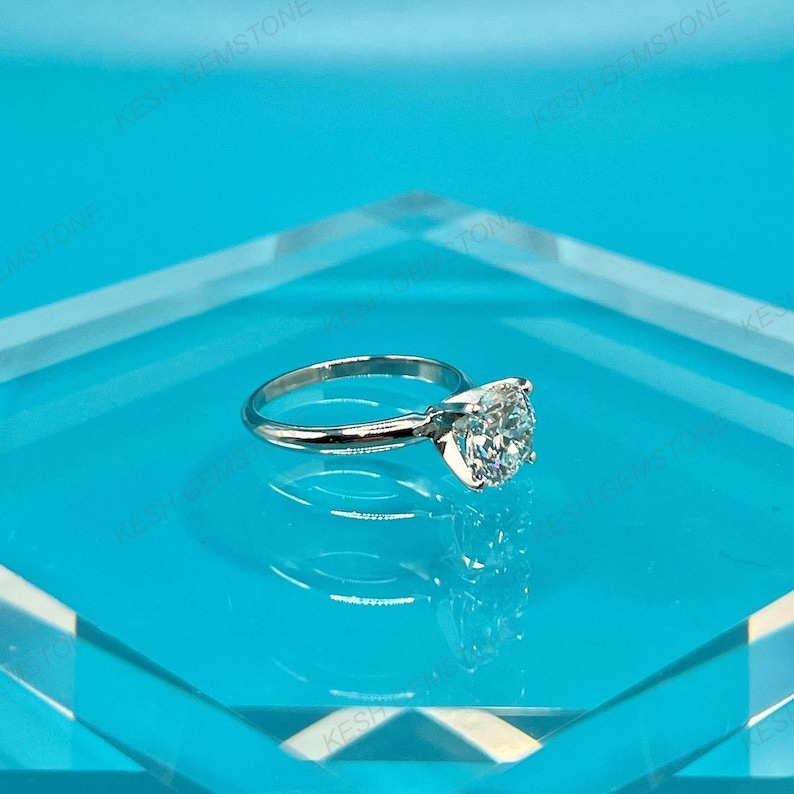Op de afbeelding: Een zilveren verlovingsring met een grote, ronde, heldere edelsteen. De ring ligt op een helderblauwe ondergrond, met de merknaam "KESH GEMSTONE" zichtbaar op de achtergrond.